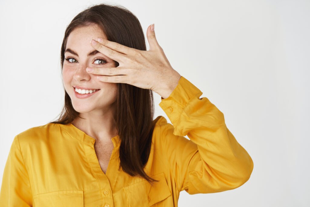 Beauty. Close-up of happy young woman looking through fingers and smiling, standing over white background